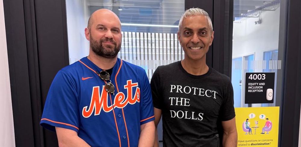 Dr.s Seth Abrutyn (L) and Amin Ghaziani (R) stand in front of the Equity and Inclusion office at the University of British Columbia. Ghaziani wears a black shirt that reads, in white lettering, PROTECT THE DOLLS.