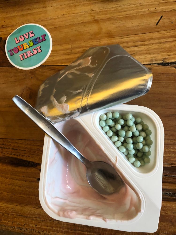 Joghurtpackung, aus der der Joghurt gegessen wurde und im Knickohr noch alle grünen Kugeln sind.Oben links ein Sticker mit den Worten „Love yourself first“. Alles so auf dem Esstisch vorgefunden.