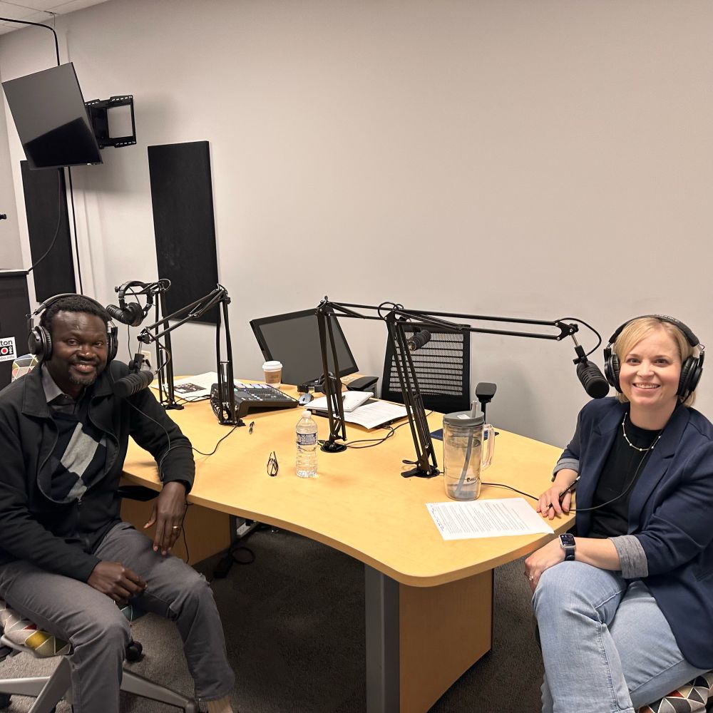 Man and woman smiling, sitting in a podcast recording studio with headphones on. 