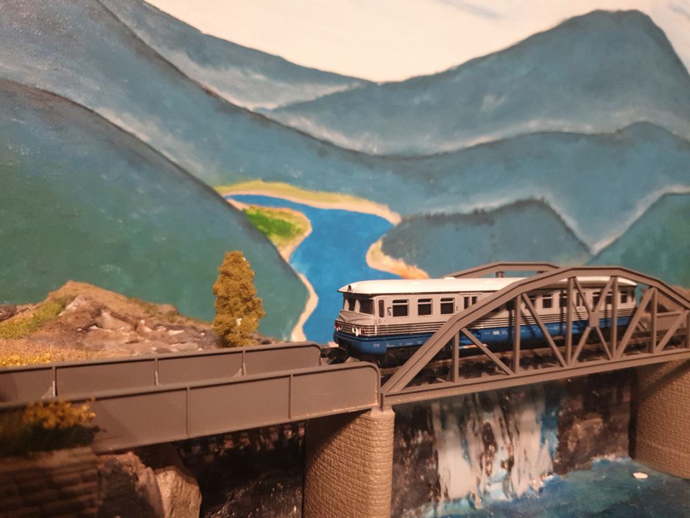 Dieseltriebwagen der Modellbahn steht auf einem eisernen Viadukt welches über einen Wasserfall führt. Im Hintergrund hohe Berge und ein Flusstal.