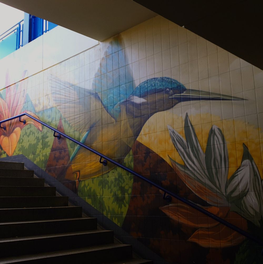 Treppe am Bahnhof führt nach oben. Die geflieste Wand ist mit einer großen farbenfrohen Malerei versehen. Ein blauer Vogel fliegt über Wiesen und Blumen.