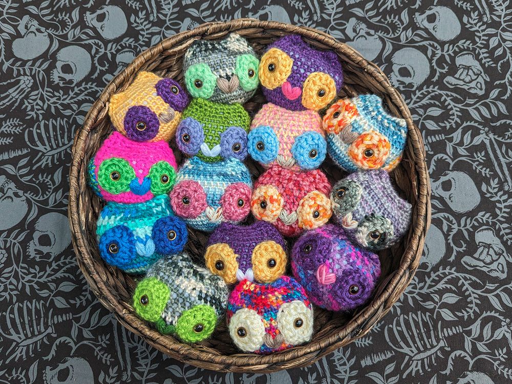 Assortment of crocheted amigurumi pride owls nestled in a low wicker basket shot from overhead. Owls include nonbinary, genderqueer, lesbian, sapphic lesbian flag, asexual, polysexual, pansexual, aromantic, aroace, intersex, bisexual, trans, agender, and rainbow pride flag owls.