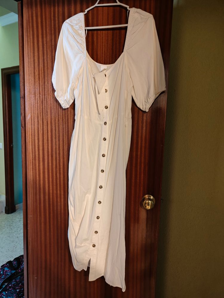 Vestido beige o rosita muy claro largo, con botones por delante y mangas abullonadas. Cuelga de una percha blanca enganchada a la puerta de madera oscura de mi dormitorio. Se ve la pared verde. 