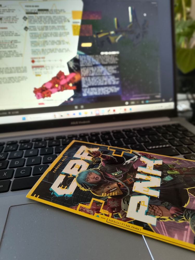 Um notebook com uma tela um pouco desfocada com um material de RPG e no primeiro plano, sobre o teclado, a parte de cima do panfleto de regras do CBR+PNK