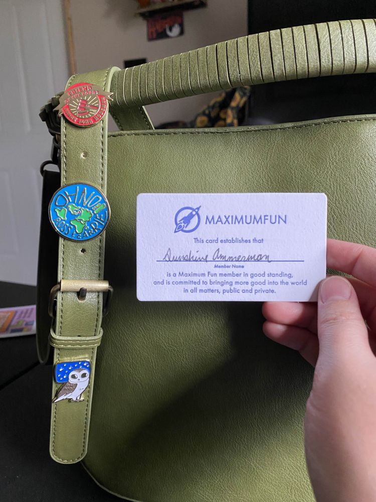 A closeup of the green bag strap and pins shows two Oh No Ross and Carrie pins and a Judge John Hodgeman pin and the Maximum Fun membership card which reads "MAXIMUMFUN This card establishes that (handwritten - sunshine ammerman) member name is a Maximum Fun member in good standing, and is committed to bringing more good into the world in all matters, public and private."