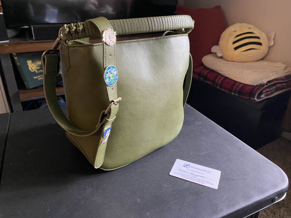 A green bag with three collectible maximum fun pins in the strap sits on a black table next to a maximum fun membership card. There is a bumblebee squishmallow in the background. 