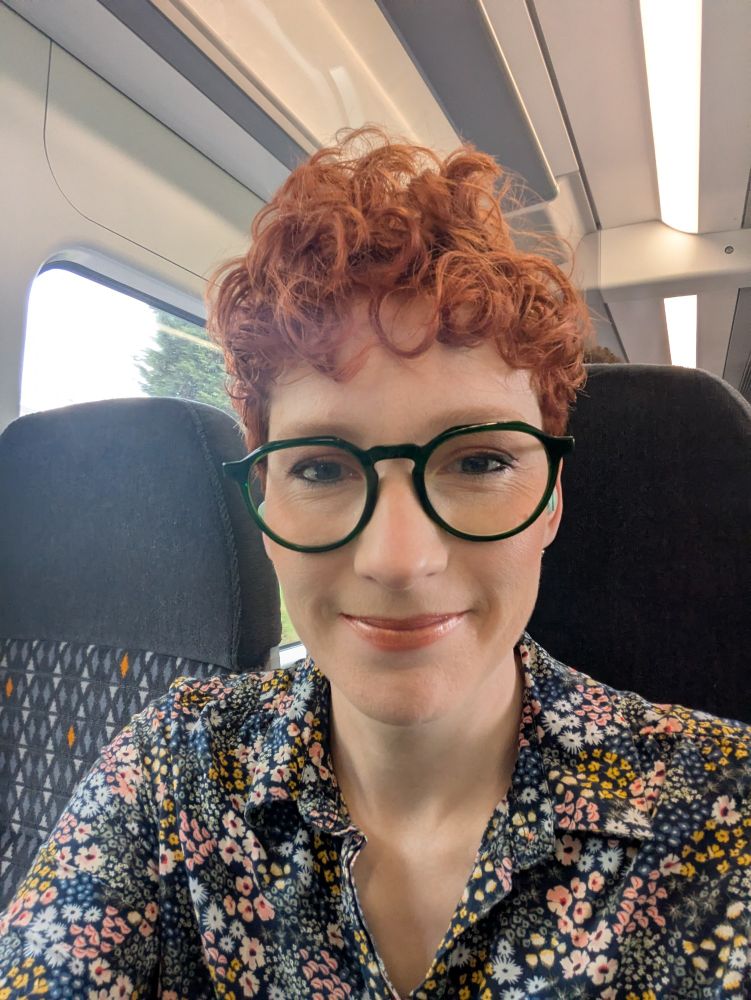 Katie, a red-headed person wearing a flowery shirt and green framed glasses, is sitting on a train. They are smiling at the camera. 