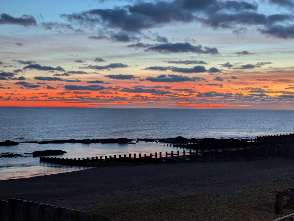Sunset over the sea. The sky is light blue fading to a bright red at the horizon. Dark clouds stream the sky. In the foreground the beach is a dark silhouette, the upright pillars of a ground like a fence across the beach. 