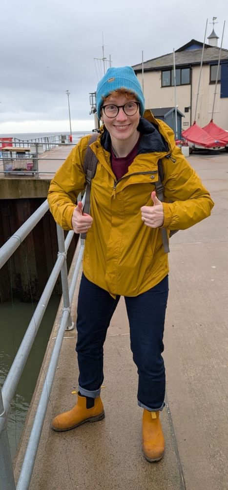 Katie in a yellow jacket and shoes and blue hat, posing with two thumbs up on the jetty at Aberdovey 
