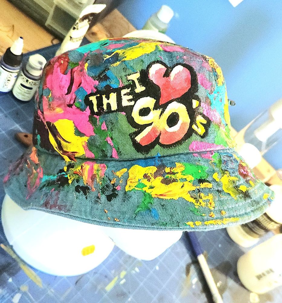 Photograph close up of a painted denim bucket hat. The hat is covered with various colorful paint slotches with the center of the hat having lettering that reads "I Love (Heart) the 90's"