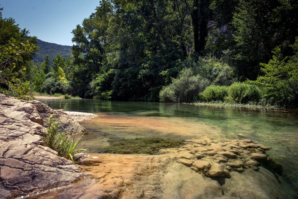 The waters of the Verdouble River flow slow and clear,
laying bare the textures of the rocky bed with an honesty only nature knows.
There is no noise, only the whisper of leaves
and the soft echo of water caressing stone.
The banks, draped in wild vegetation, invite rest, and let time dissolve.
In the distance, the hills wear a deep green,
like silent guardians of this untouched corner.
A place that asks for nothing, only your presence.
And in that stillness… beauty.