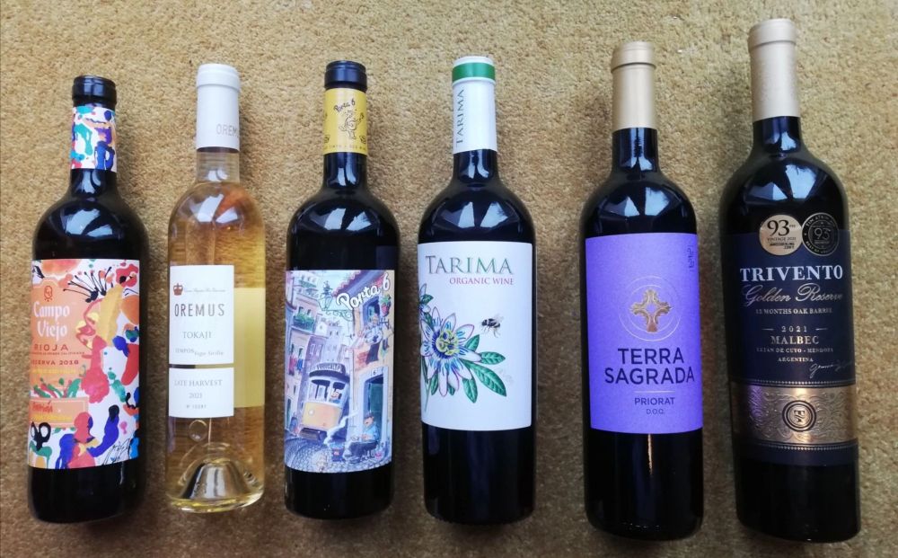 Photograph of six unopened wines bottles, five reds and one white, on a beige background.