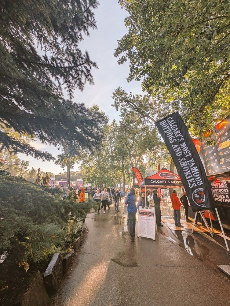 Blue sky poking through after a heavy rain. Trees on either side of a pathway, with folk music festival stage in the back. Food vendors along right hand side of pathway. 