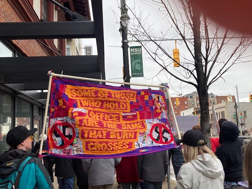 A multi-colored, quilted sign that says "Some of those who hold offices are the same that burn crosses." It is being held by two people, including the badass who took 80 hours to make this.
