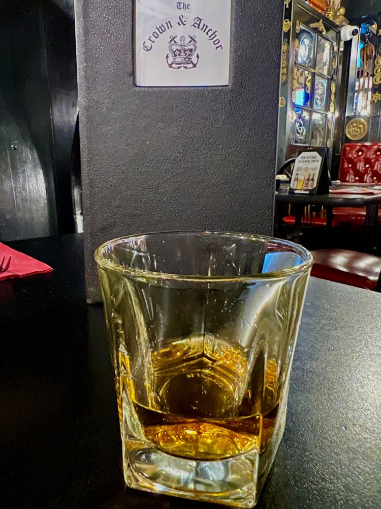 A rocks glass with whiskey in it, with a Crown & Anchor pub menu behind it. 