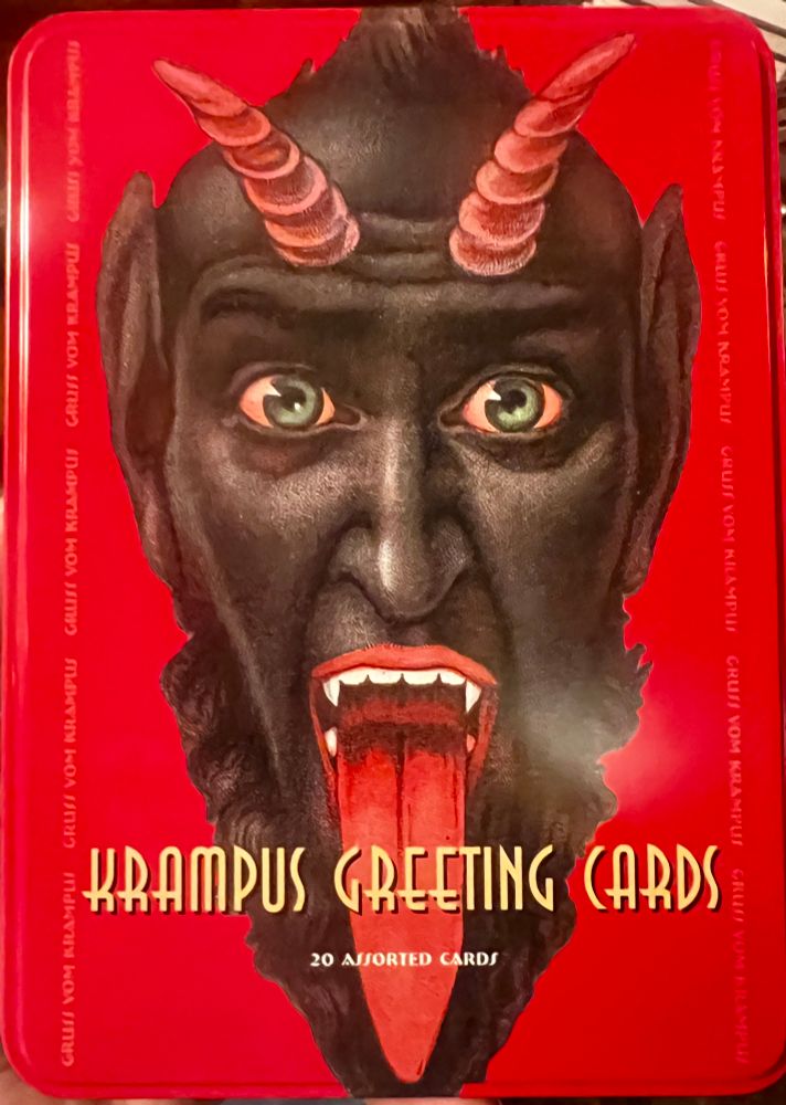 A photograph of a tin featuring a demon with long pointed ears, dark skin, pink horns coming out of its forehead, green eyes, fangs, and a very long tongue coming out of his mouth. The caption says KRAMPUS GREETING CARDS. 