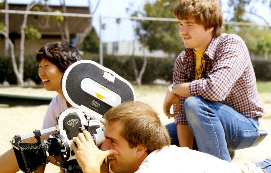 Me squatting down, wearing blue jeans and a red & black checked shirt over an Eclair NPR T-shirt, with shaggy hair and a silly little mustache. My friend Paul is looking through the eyepiece of an Arriflex 16mm camera. Carolyn, the director of the shoot, is behind him smiling. 
