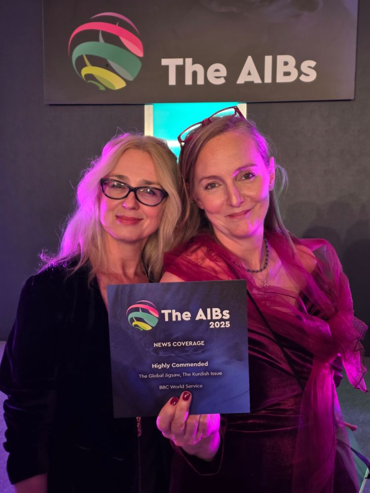 At the AIBs with our amazing presenter of The Global Jigsaw, Krassi Twigg (in black), proudly holding up our commending certificate, it front of the AIBs board - in front of a dark backgroubd with the letters logo of the AIBs two women in formal evening clothes smiling, looking into the camera