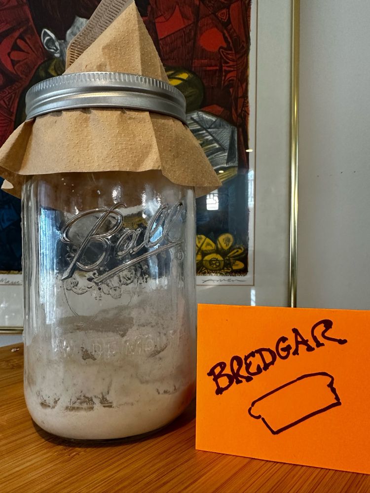 A mason jar with sourdough starter in it, with a handwritten card next to it that says Bredgar and a poorly drawn load of bread 
