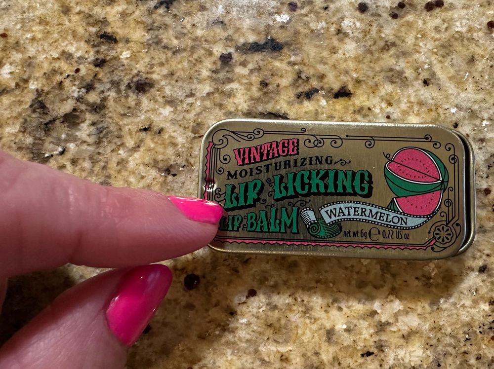 A tin of “vintage” moisturing lip licking lip balm, watermelon “flavor” (scent) being pointed to by fingers painted the same color as the watermelon  drawing depiction on the tin