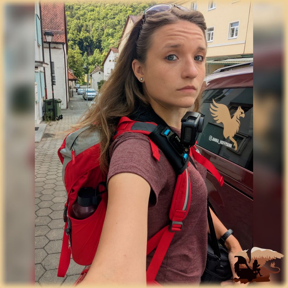 Selfie von mir (Frau Ende 20, dunkelblonde Haare, Sonnenbrille auf dem Kopf, mit weinrotem T-Shirt, dem rot-grauen Wanderrucksack auf dem Rücken und am rechten, der Kamera zugewandten Schultergurt einer Akaso Actioncam. Im Hintergrund zu sehen mein rotes Auto mit Logo und Insta-Handle. Mein Blick ist sehr konzentriert bis skeptisch. 