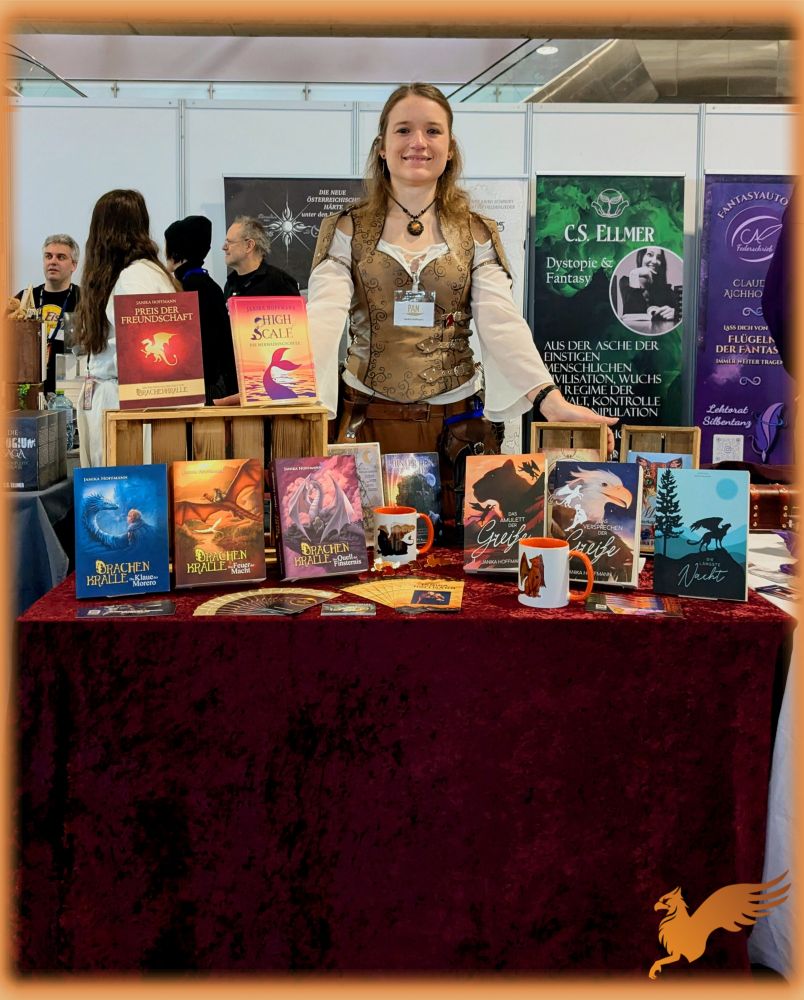 Foto von mir in mittelalterlich inspiriertem Fantasyoutfit hinter meinem Tisch mit roter Pannesamtdecke. Darauf befinden sich meine Fantasyromane, einige Kurzgeschichtensammlungen, Tassen und weiterer Merch. 