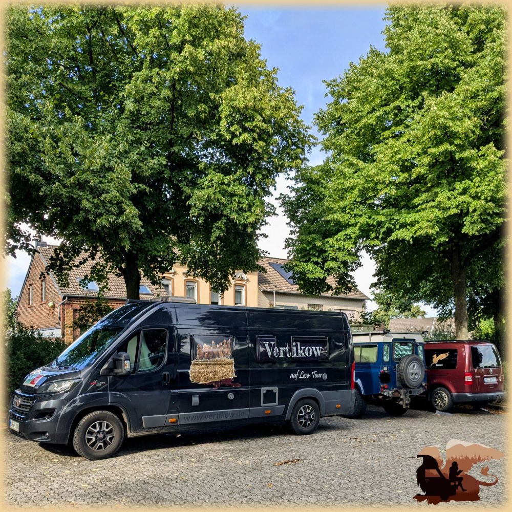 Drei Camper stehen nebeneinander in einer durch Bäume und Hecke begrenzten, gepflastertrn Parzelle: ein 6,50 Kastenwagen in schwarz mit Aufdruck "Vertikow auf Lesereise", daneben ein dunkelblaur Defender mit weißem Dach und daneben auf Auffahrkeilen ein Caddy in rot-metallic mit goldenem Greifenlogo und Insta-Namen @greivan.tv als Beklebung. 