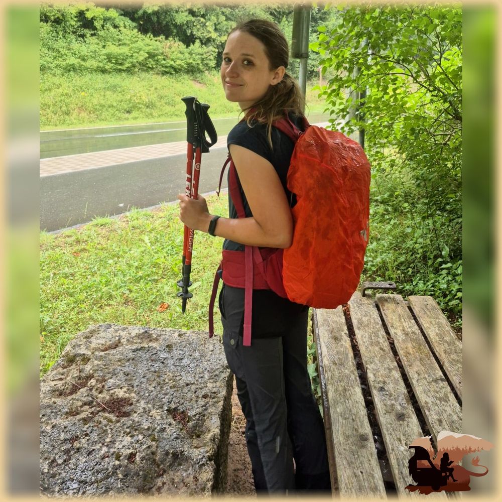 Foto von mir (Frau Ende 20, dunkelblonder Pferdeschwanz, dunkle Wanderhose und T-Shirt) zwischen einer Holzbank und einem steintisch am Rand einer Straße unter einem Baum. Es regnet, ich bin ebenfalls nass. Ich drehe den Kopf halb, um in die Kamera zu grinsen. Mit der linken Hand halte ich meine roten Wanderstöcke hoch. Auf dem Rücken habe ich einen Wanderrucksack, der in einen orangefarbenen Regenschutz mit Vaude-Logo drauf eingehüllt ist. Der Regenschutz ist ebenfalls schon ziemlich nass. 