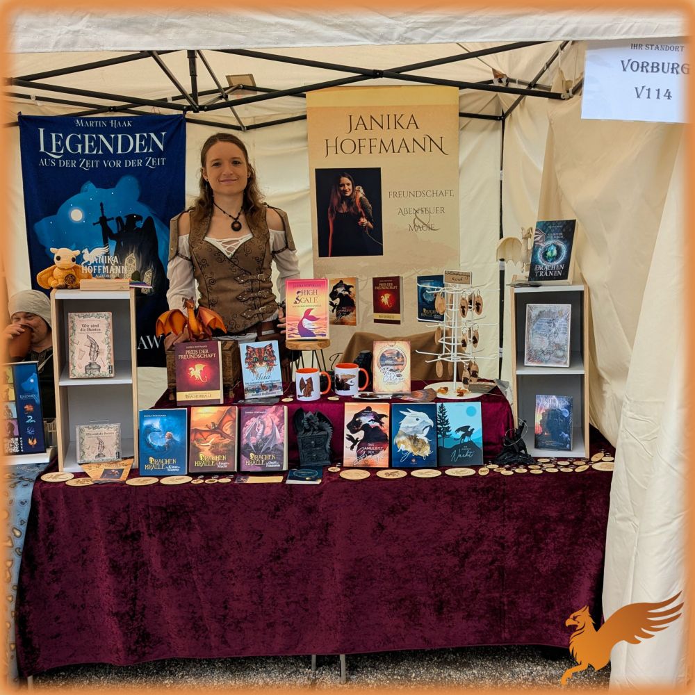Foto eines Stands mit Büchern, Tassen und Schlüsselanhängern unter einem cremefarbenen Faltpavillondach. Ich stehe hinter dem Stand: Frau Ende 20,lange,dunkelblonde Haare, weiße Bluse, Lederwams und eine Kette mit orangefarbenem Anhänger. 
