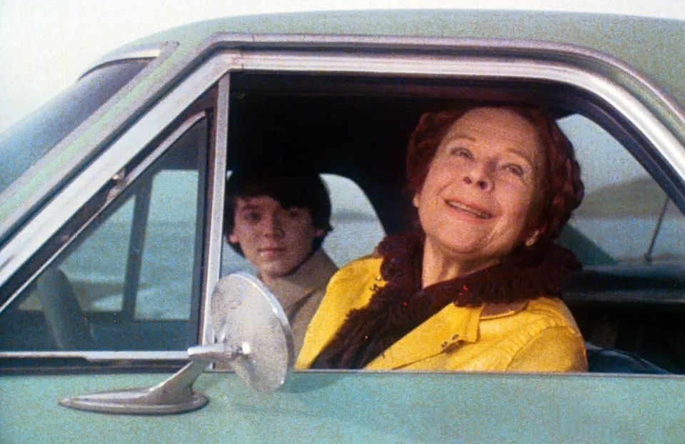 Picture from the movie Harold and Maude. Maude is driving a blue car with a smile on her face; Harold is in the passenger seat.