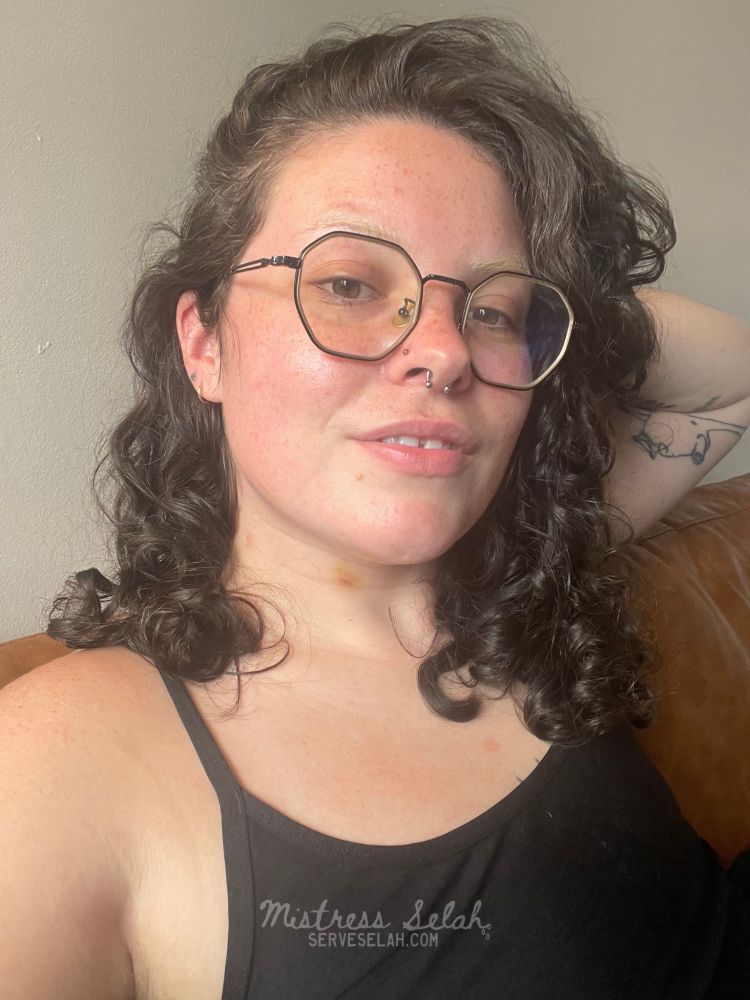 Mistress Selah with freshly washed curly hair and a yellowing hickey on her neck. She smiles softly at the camera 