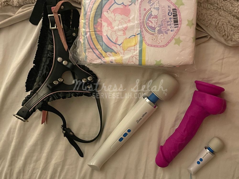 An assortment of toys on a cream colored bed that Mommy used on her baby: a beautiful leather strap harness with pink leather ruffles, a diaper adorned with unicorns and stars, a big magic wand and a mini magic wand, and Mommy’s favorite pink cock