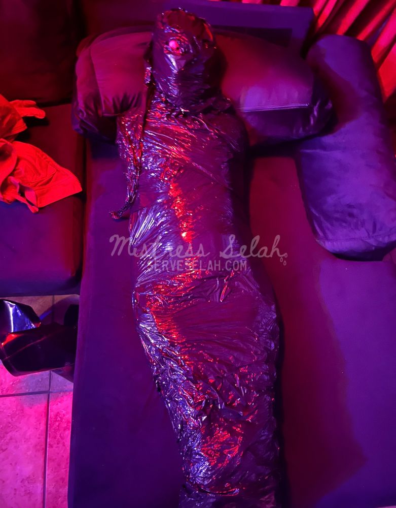 Selah's sub entirely encased in black plastic wrap and tape, lovingly laid out on a couch and bathed in red light