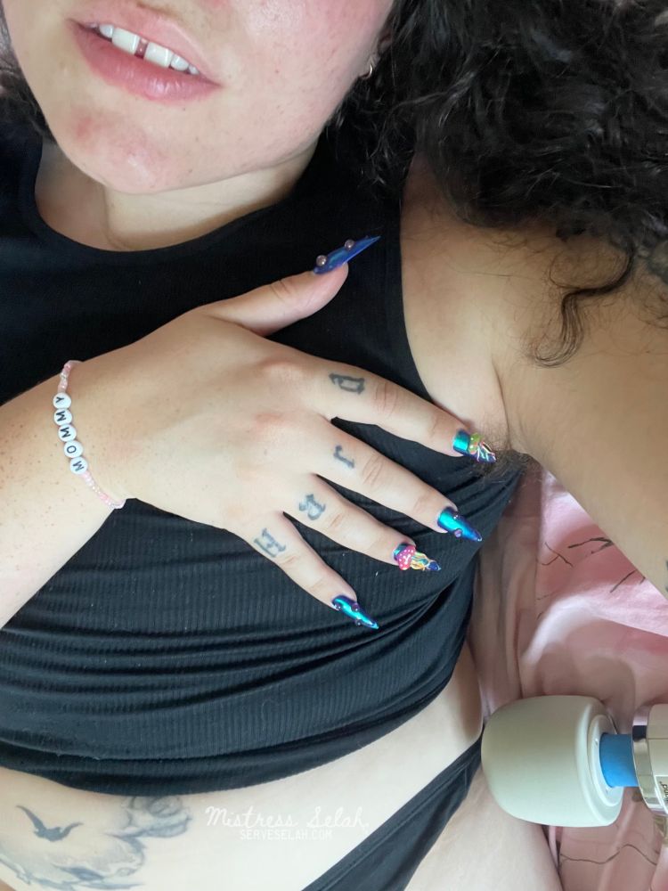 A woman in simple black thong and black top spread over pink bedsheets. One hand with long, blue fingernails lays over her chest. Her knuckles are tattooed with the word “hard” and on her wrist she wears a pink bracelet that says “Mommy”. She has sweaty, hairy armpits and curly brown hair. The head of a hitachi can be seen in the corner of the photo