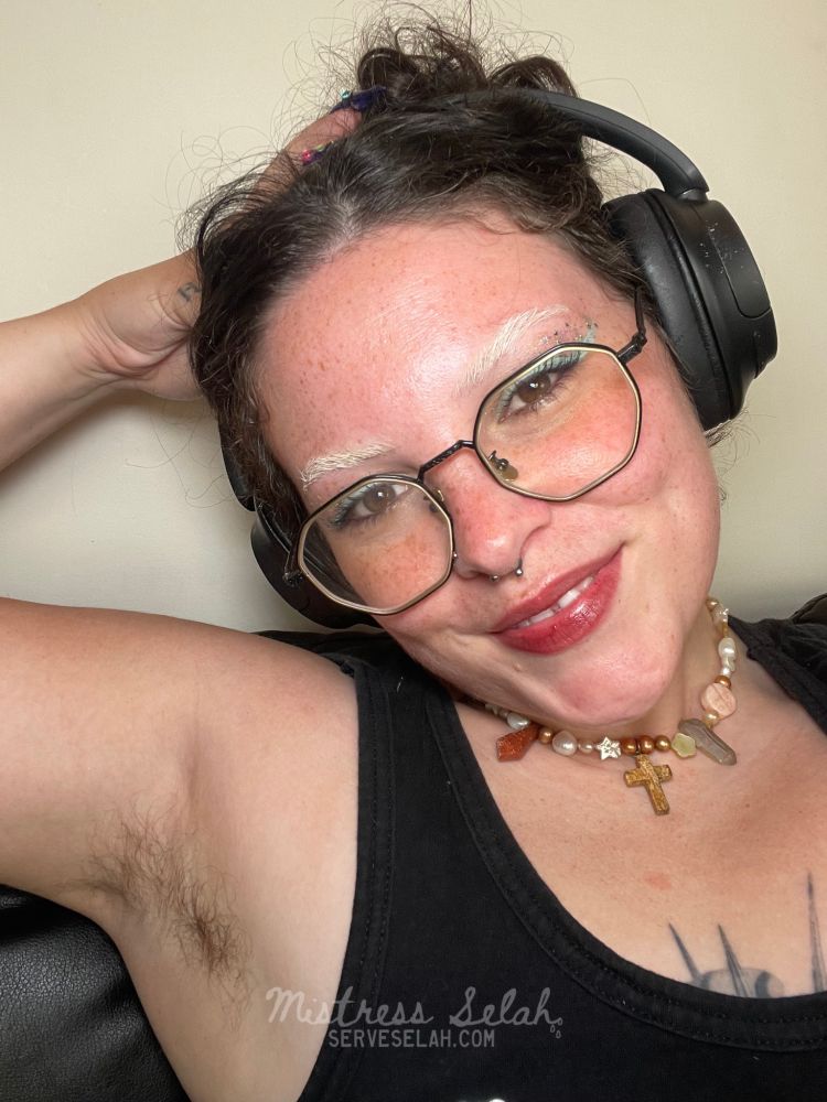 Selfie of Mistress Selah in casual wear smiling devilishly. She has messy hair in an updo and funky makeup on. Her arm holds her head up revealing her hairy armpit 