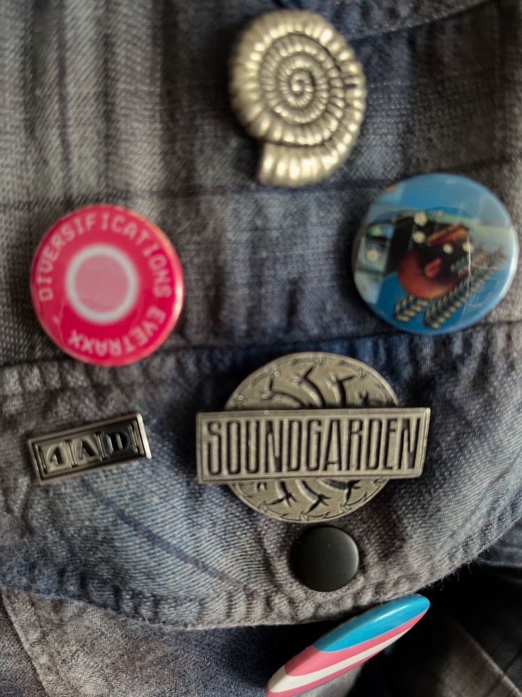 Badges for Nicola Griffith’s Ammonite, Pat Cadigan’s Synners, Soundgarden, @, and 4AD on a plaid shirt 