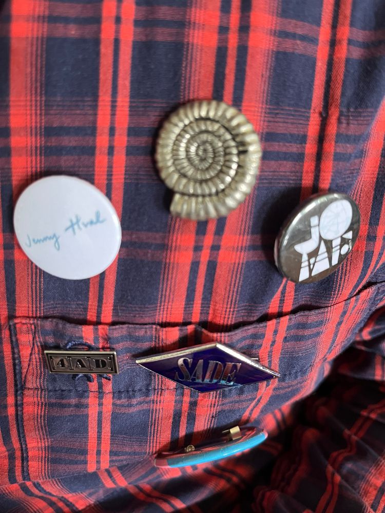 Badges for Nicola Griffith’s Ammonite, Jenny Hval, Sade, JOMF and 4AD on a plaid shirt 