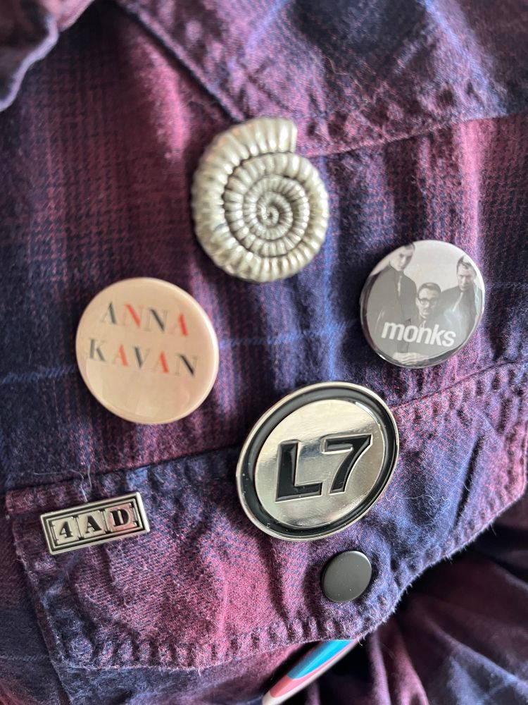 Badges for L7, the Monks, Nicola Griffith’s Ammonite, Anna Kavan, and 4AD records on a plaid shirt