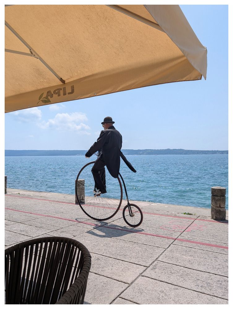 Auf einer Strandpromenade am Meer fährt ein älterer Herr mit Smoking und Hut auf einem historischen Hochrad. Im Vordergrund ein Stuhl und ein Teil eines Sonnenschirms von einem Eiscafe. 