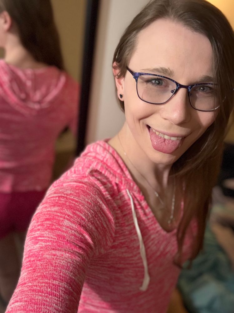Katie, in full playful mode, sticks her tongue out with a cheeky grin. Wearing her pink hoodie and purple-framed glasses, her fun-loving personality shines through. Her long hair cascades down one shoulder, and the mirror in the background shows her pink shorts, adding to her effortlessly cute and flirty style.