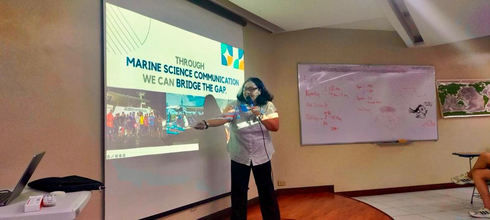 Picture of a woman conducting a talk on marine science communication. Projected behind her, the slide reads “through marine science communication we can bridge the gap”