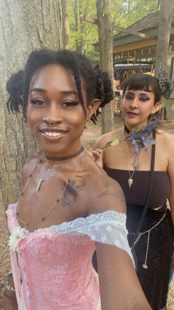 A selfie of me and my roommate who is standing out from behind my shoulder. I’m wearing a pink corset and white lace off shoulder sleeve cuffs and my roommate is wearing a black tune top and ornately designed black shawl skirt and waist chain. We’re dressed as fairies with pointy ears and glitter makeup, outdoors with tall trees in the background. My roommate holds up a purple lily wand under their chin. #blackfae #blerd 