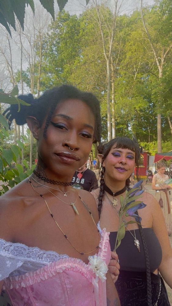 Another selfie of the roommate and I outdoors in our fairie outfits. You can see ran faire vendor carts and patrons in the background. We’re smirking and squinting. 