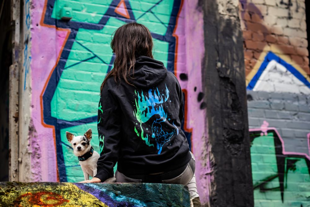 photo of a woman with a chihuahua wearing a black hoodie with an aurora fox backprint. In the background is a graffiti covered wall