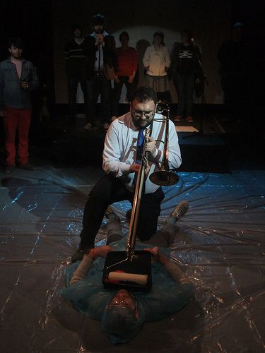 Ben being hazed by Nathan playing paint roller trombone over him, as the assembled Board members watch from the back of the stage