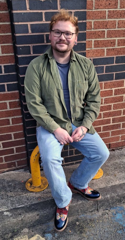 Me sat outside in a green shirt and blue jeans, with super cool fancy shoes. I have my glasses on with a scruffy beard and ginger hair.