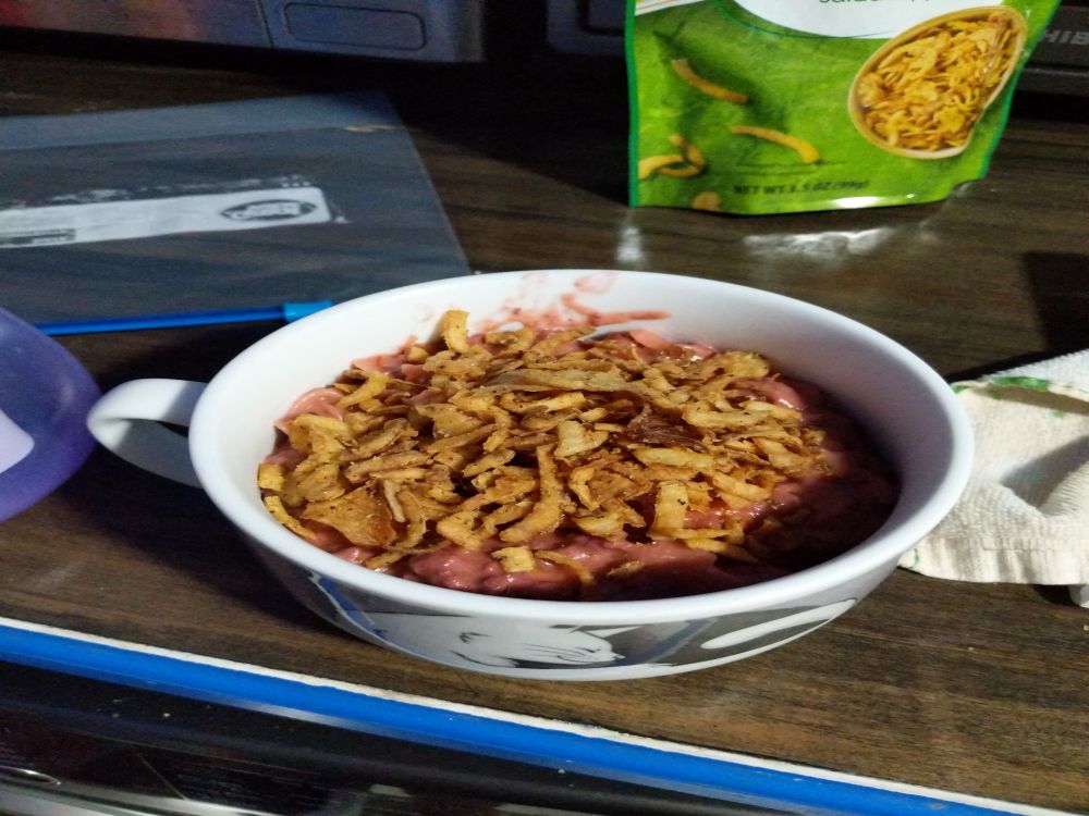 The previous mixture in a bowl with two cats hugging, one black and one white. The mixture is now a darker pink and has crispy onions on top, the bag for them can be seen in the background 