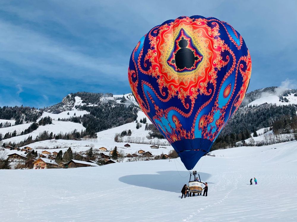 Fractal hotair balloon on a snowy field in the Swiss Alps being carried out to a road for deflation, with mountains in the background 