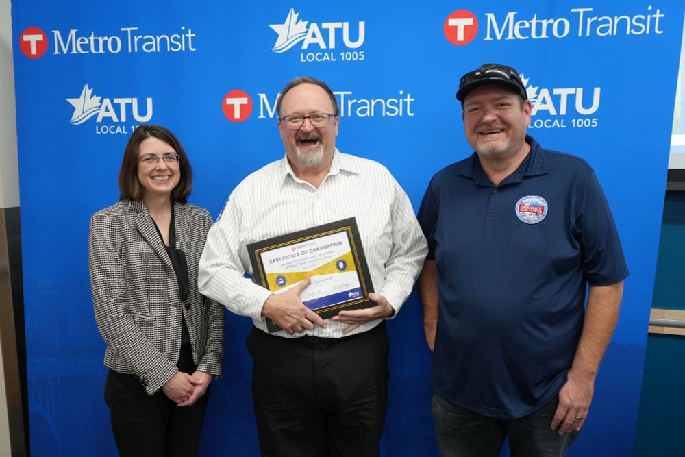 A bus operator graduate stands with their graduation certificate.