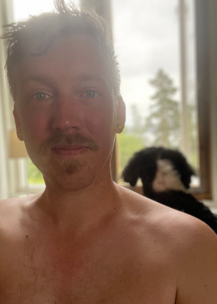 Nöjd man framför ett fönster. Mustach och bar överkropp, men typ bara axlarna som syns. Fantastisk hund som syns i bakgrunden. 